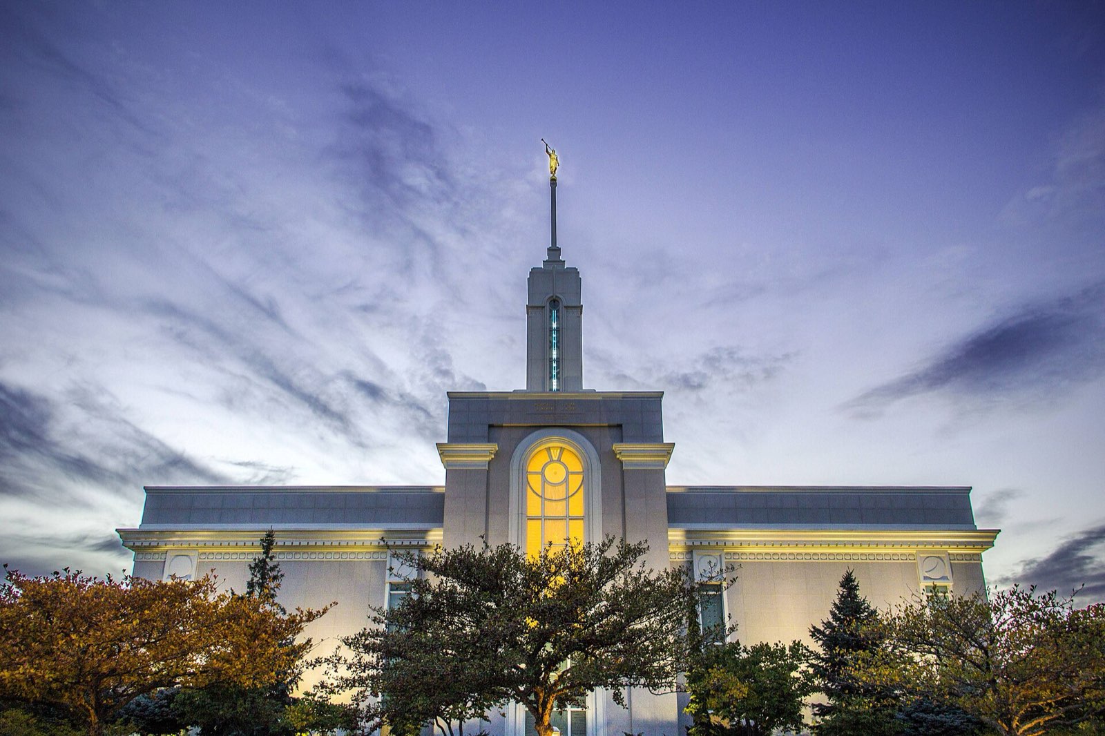 The Mount Timpanogos Utah Temple in American Fork at dusk, its lit center window and spire rising above autumn trees